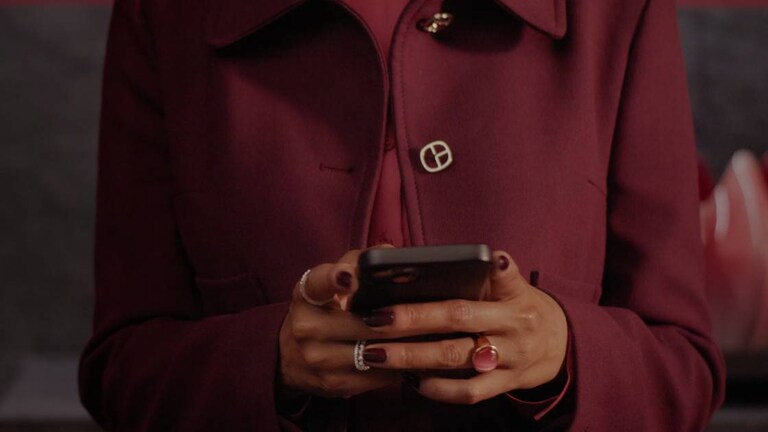 Close-up van een persoon in een dieprode jas met gelakte nagels en ringen, die een mobiele telefoon gebruikt