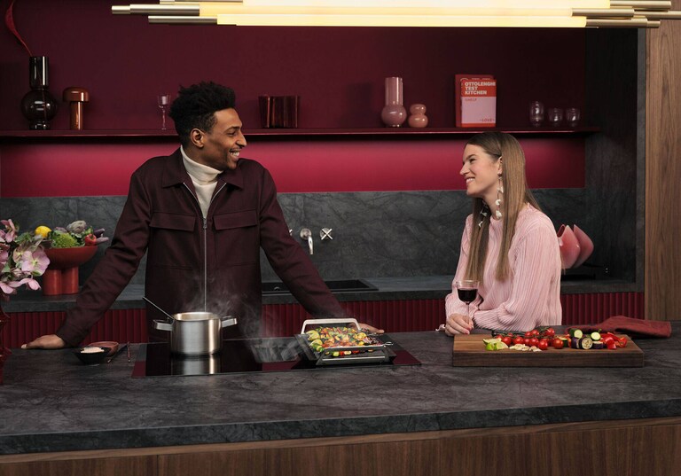 Man en vrouw in keuken aan het praten in een keuken waar een ingebouwde inductiekookplaat ligt waar ze voedsel op aan het bereiden zijn.