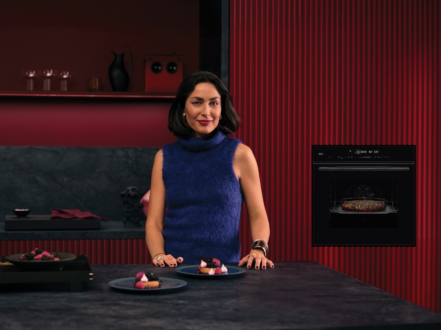 Vrouw in blauw shirt in een bordeaux rode keuken met in de achtergrond een ingebouwde zwarte AEG heteluchtoven met magnetronfunctie en een aanrecht waar borden klaar staan met voedsel erop.